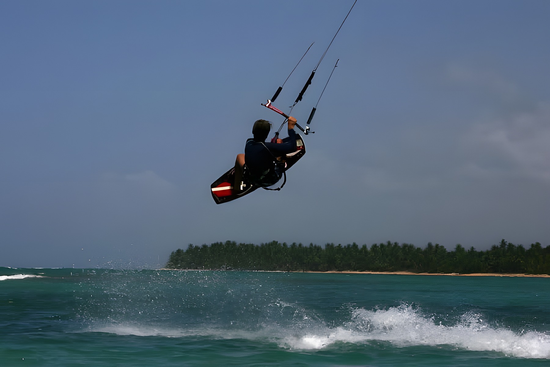 Teenage Hugo Beaumont mid-air on a kiteboard jump above a palm-lined bay